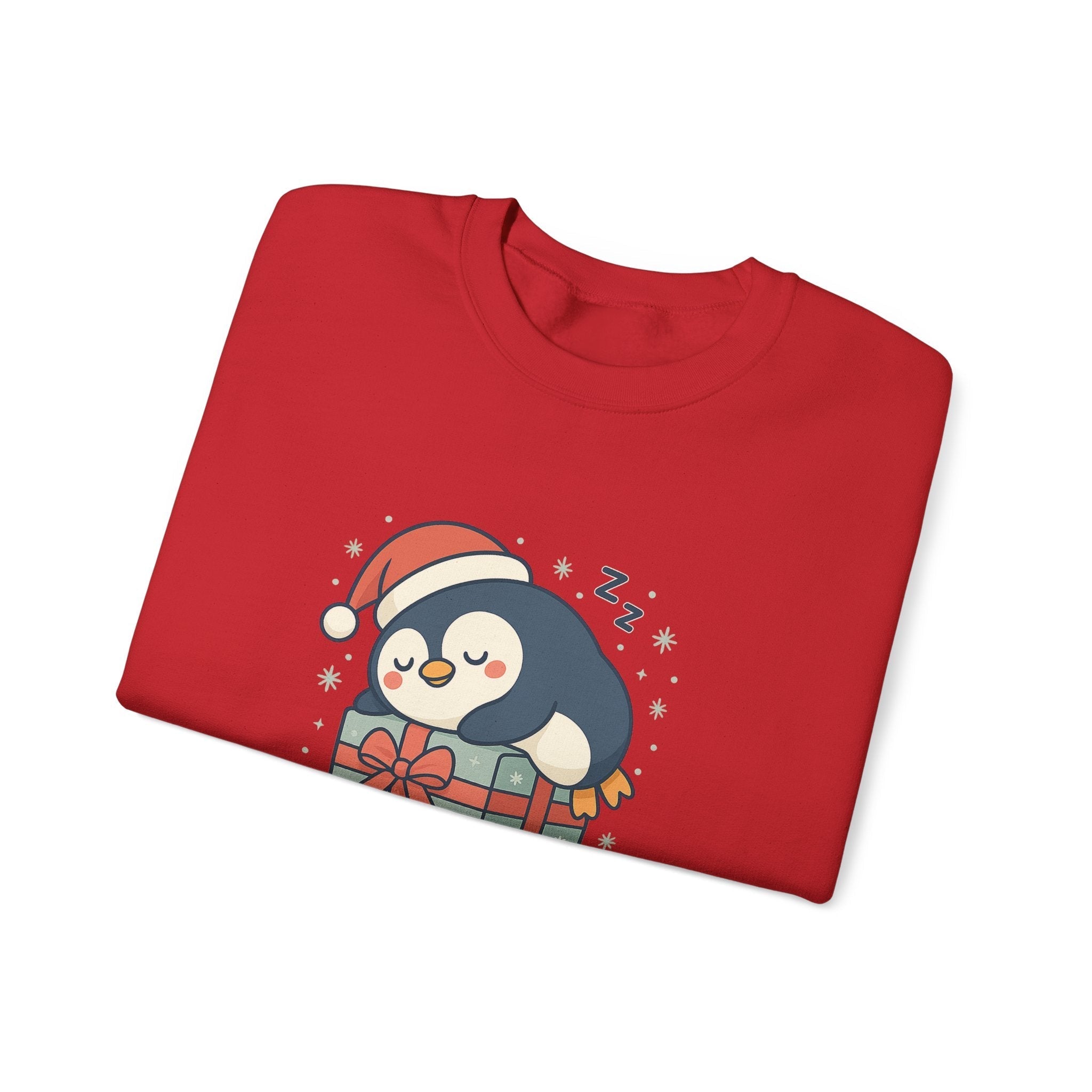 Folded red Christmas sweatshirt with cute chubby penguin sleeping on a present, Santa hat design, cozy winter holiday pullover for adults by DJ’s Giggle Wear.